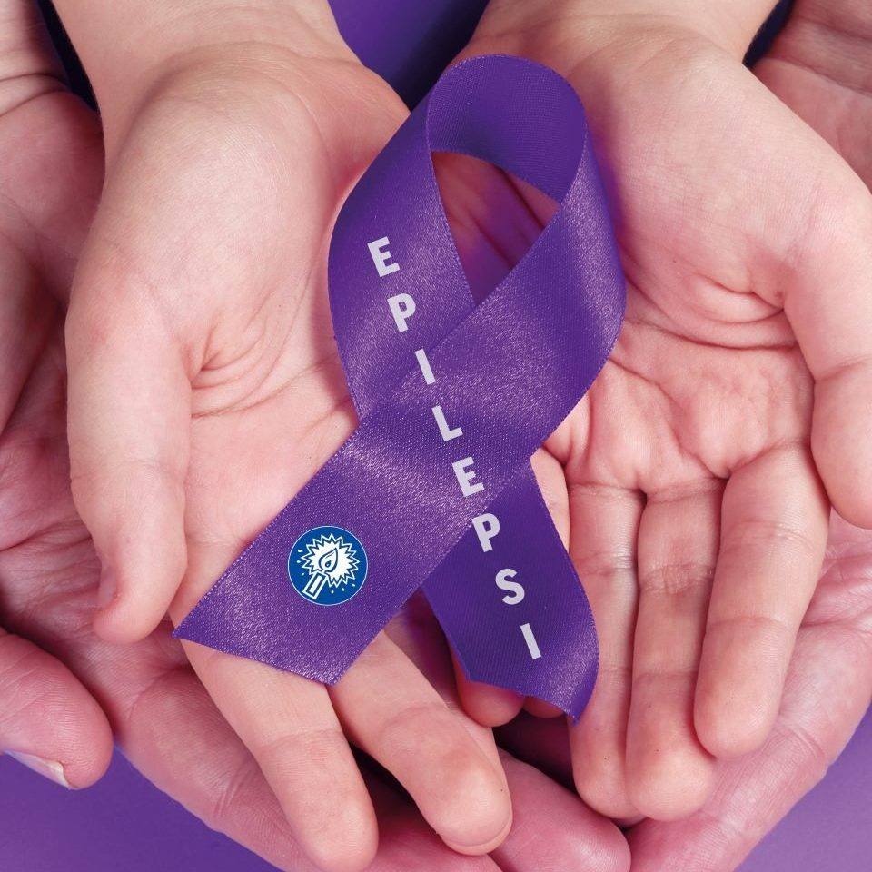 ett barns händer i en vuxenshänder med handflatorna upp och i dem ett lila band med epilepsiförbundets logotyp det brinnande ljuset och texten epilepsi
